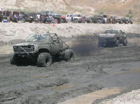 1985 Ford Bronco - Two heavily modified trucks are stuck in thick mud, with one being towed by the other as onlookers watch from a sandy embankment.
