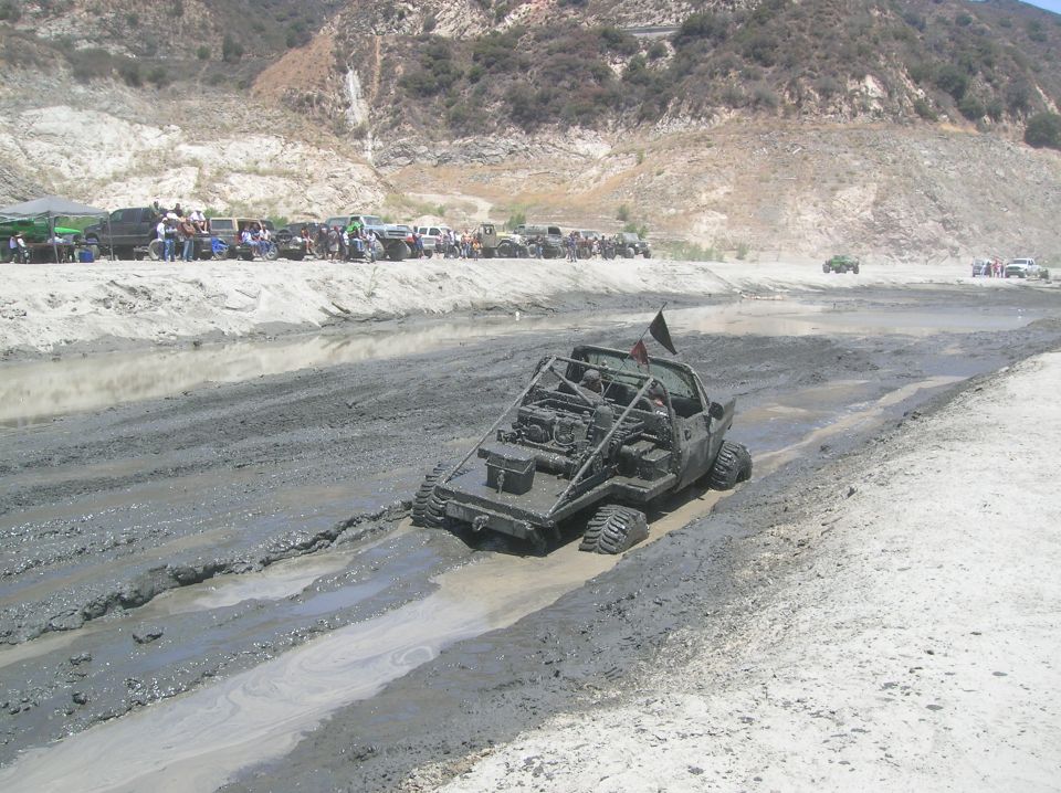 Custom buggy with exposed engine and roll cage driving through mud pit; other vehicles and spectators in background
