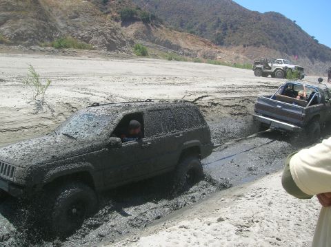 1985 Ford Bronco - Two off-road vehicles are stuck in thick mud, with one being pulled out by the other.