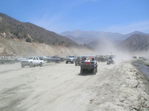 1985 Ford Bronco - A group of off-road vehicles are gathered in a dusty, mountainous terrain, with some driving and others parked.