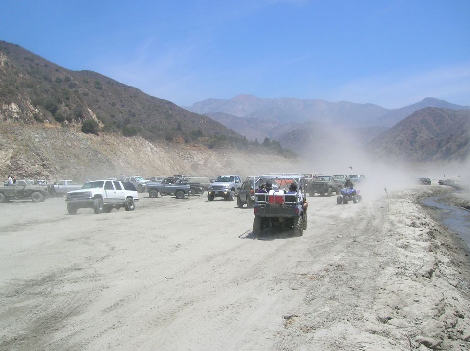 Dusty scene with many trucks and ATVs parked and driving near a mountain