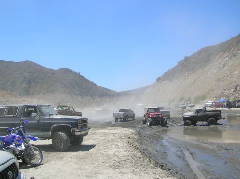 1985 Ford Bronco - Off-road vehicles are parked and driving in a muddy, dusty terrain under a clear blue sky.