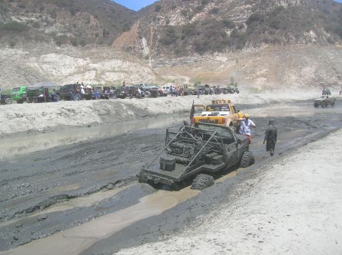 1985 Ford Bronco - Several off-road vehicles are navigating a muddy track with onlookers gathered on the side.