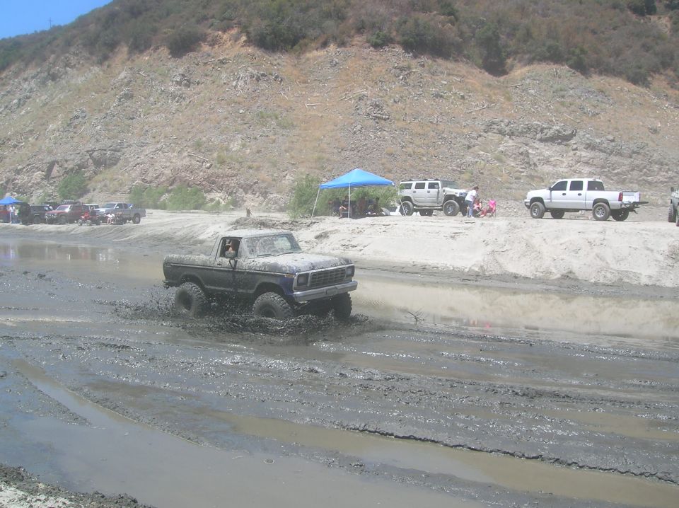 Mud-splattered dark blue Ford pickup truck driving through a muddy track, front view