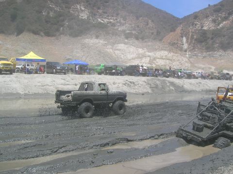 1985 Ford Bronco - A lifted pickup truck navigates a muddy track during an off-road event with spectators in the background.