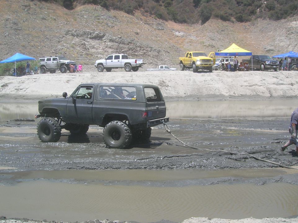 modified SUV with large tires stuck in mud, being pulled by a rope. Other trucks and tents are visible on the bank