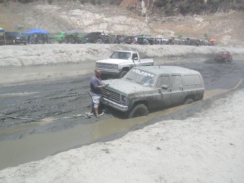 1985 Ford Bronco - A man attempts to free a mud-covered Chevrolet Suburban from a muddy ditch while other vehicles wait in the background.