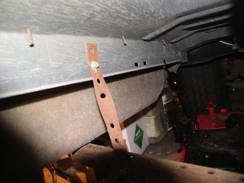 Underbody of a vehicle showing a rusty metal bracket attached to the frame, with a tire and red jack visible in the background
