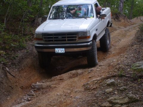 1992 Ford Bronco - A white Ford pickup truck navigates a steep, rutted dirt path in a wooded area.