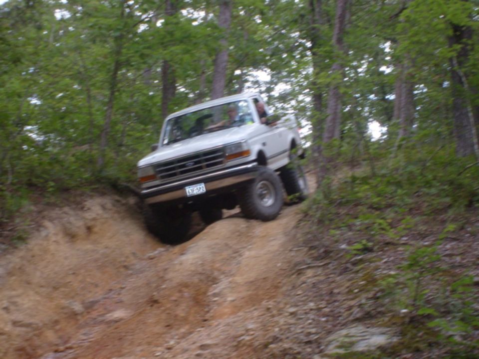 White 1992 Ford Bronco with lifted suspension driving off-road on a steep dirt incline