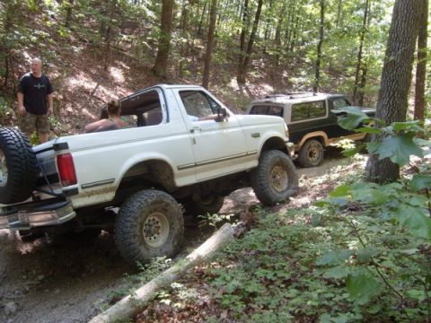 1992 Ford Bronco - Two Ford Broncos navigate a wooded trail, with the lead vehicle showing its spare tire and open-top configuration.