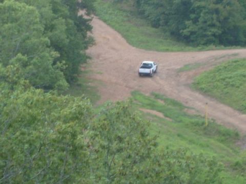 1992 Ford Bronco - A white pickup truck drives on a dirt road surrounded by trees and grass.