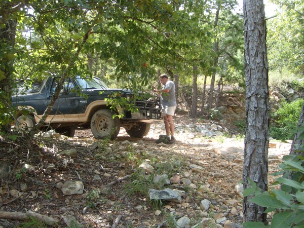 Man stands by a dark blue Ford Bronco with a tan stripe on a rocky, wooded trail. The Bronco has a brush guard
