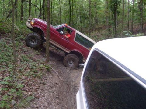 1992 Ford Bronco - An off-road SUV navigates a challenging muddy incline surrounded by trees.