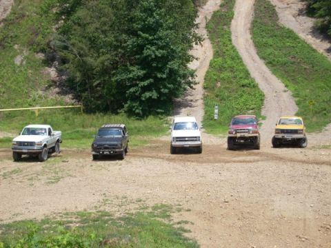 1992 Ford Bronco - Several trucks and SUVs are parked on a dirt road, with a wooded hillside in the background.