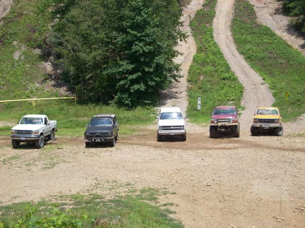 Five SUVs and trucks parked on a dirt road, with a white pickup on the left and a black SUV next to it