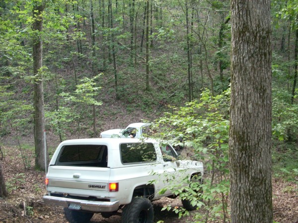 Two white SUVs driving off-road in a wooded area. The foreground SUV is a Chevrolet K5 Blazer