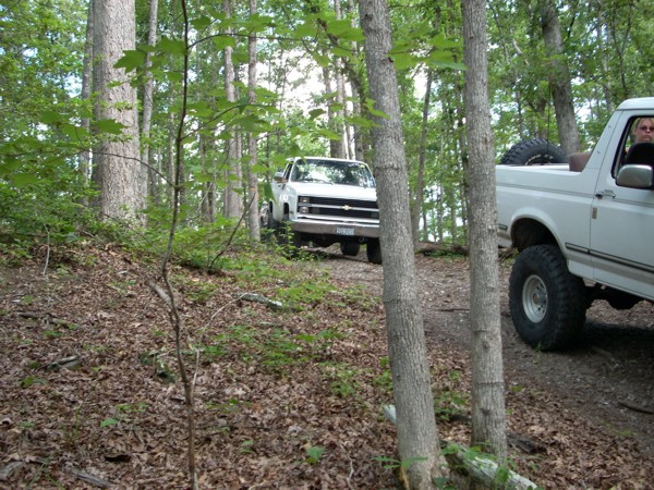 White 1992 Ford Bronco with large tires and a spare tire in the bed, parked on a dirt path in a forest