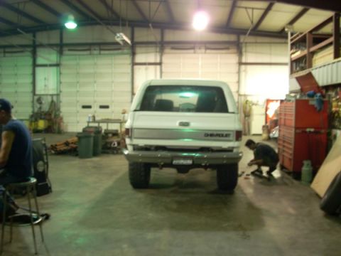 1992 Ford Bronco - A white Chevrolet SUV is parked inside a garage, with a person working on its rear bumper.
