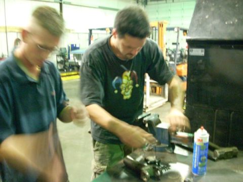 1992 Ford Bronco - Two mechanics are focused on a car part secured in a vise, with tools and a spray can nearby.