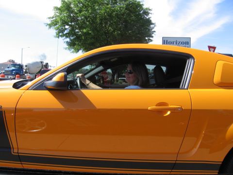 2007 Saleen S-302 - An orange coupe with black racing stripes is seen from the side, with a woman driving and a cement truck in the background.