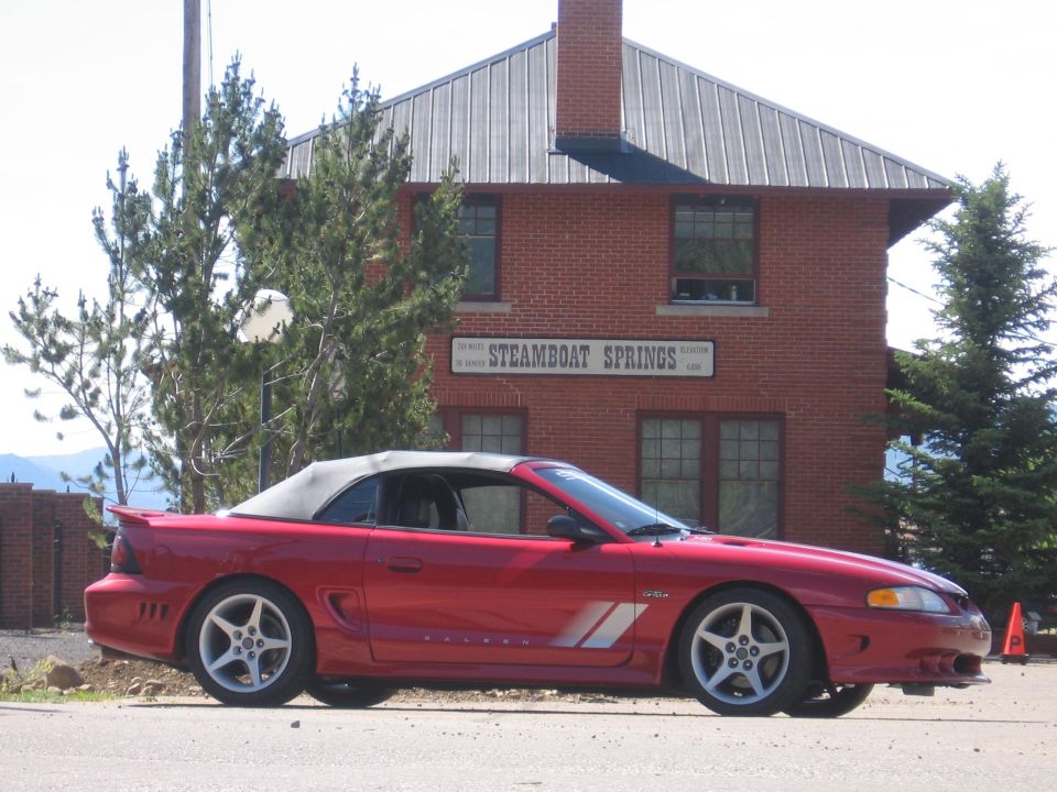 Red convertible Mustang parked on asphalt, side view. Building with 'Steamboat Springs' sign in background