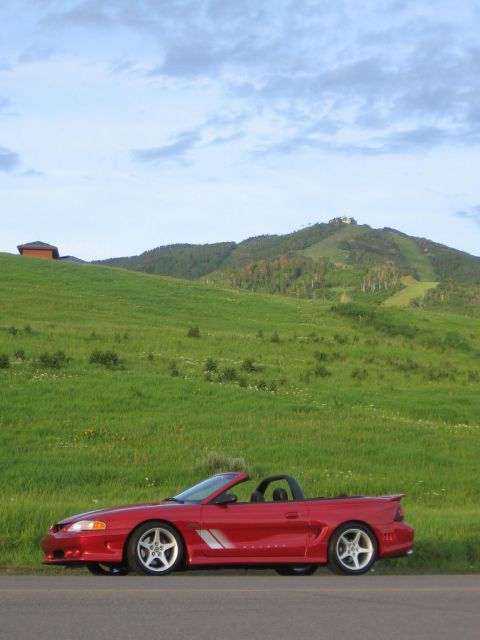 2007 Saleen S-302 - Red convertible Mustang parked on the side of a road with a grassy hill and mountains in the background.