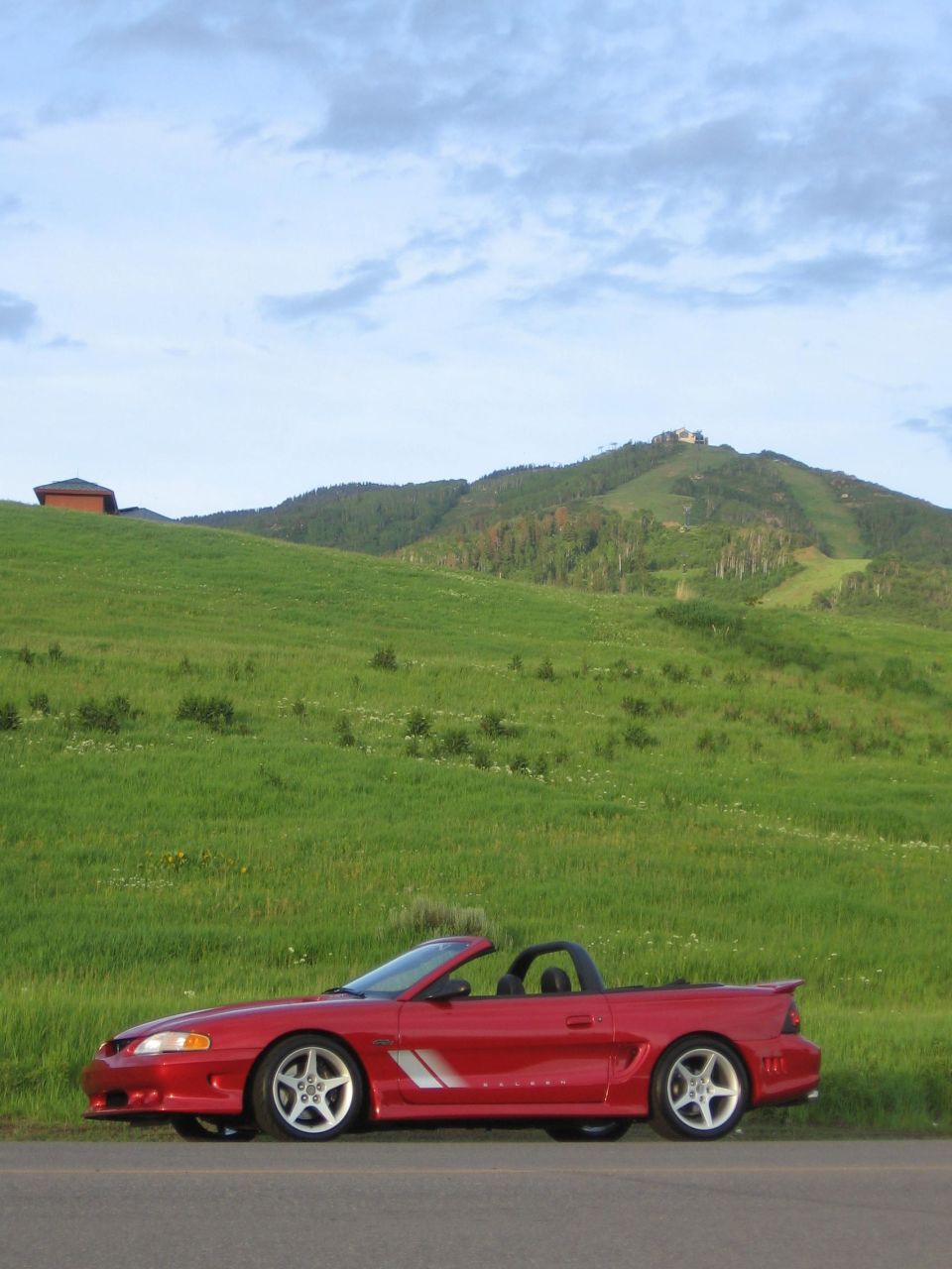 Red convertible Mustang parked on the side of a road with a grassy hill and mountains in the background