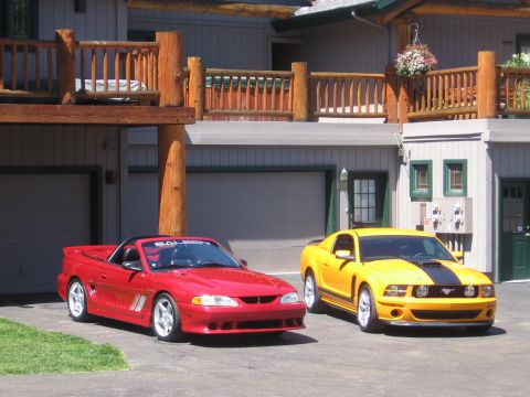 2007 Saleen S-302 - Two Ford Mustangs, a red convertible and a yellow coupe, are parked outside a house with a wooden deck.