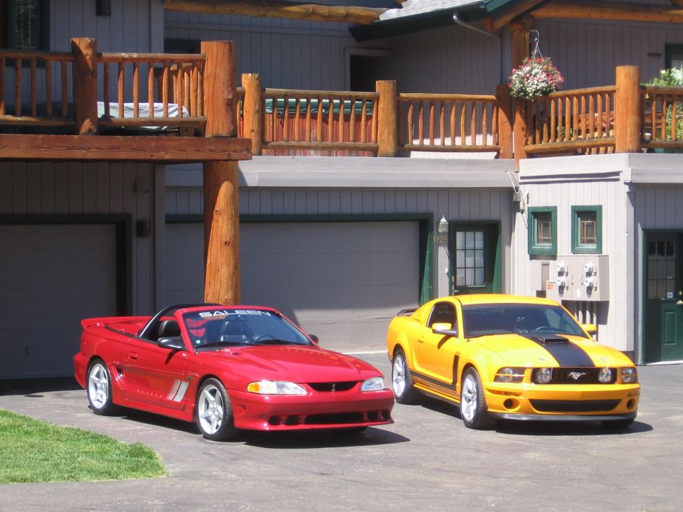 Red convertible Mustang and yellow coupe Mustang parked in front of a house