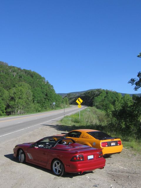2007 Saleen S-302 - Two Ford Mustangs, a red convertible and an orange coupe, are parked on the shoulder of a scenic highway.