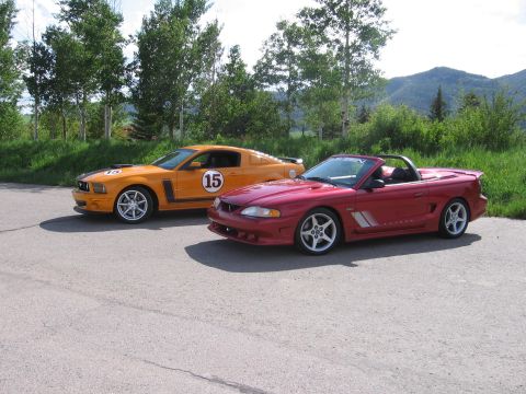 2007 Saleen S-302 - Two Ford Mustangs, one orange coupe and one red convertible, parked outdoors with trees and mountains in the background.