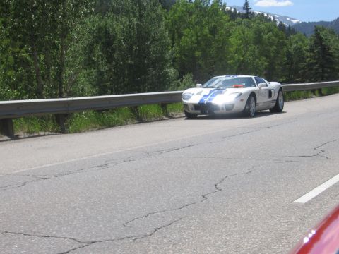 2007 Saleen S-302 - A white Ford GT with blue racing stripes drives on a highway with trees and mountains in the background.
