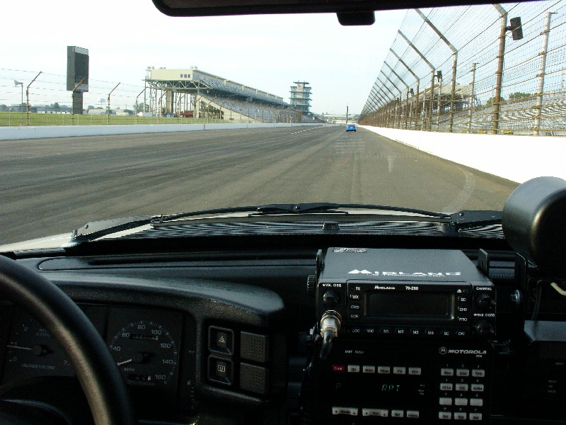 View from inside a vehicle looking out at the Indianapolis Motor Speedway track with grandstands and a distant blue car