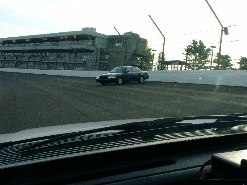 Dark sedan on a race track, viewed from inside a vehicle. The sedan has a boxy body style and appears to be a police car