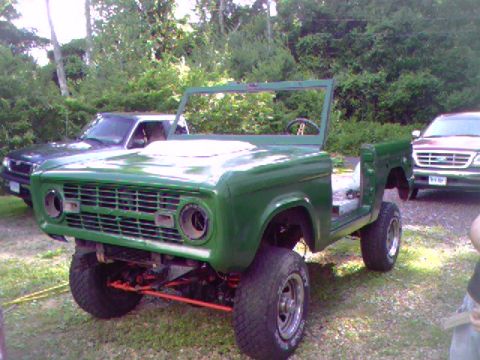 1974 Ford Bronco - This green Ford Bronco is missing its top and doors, sitting in a grassy area with other vehicles nearby.
