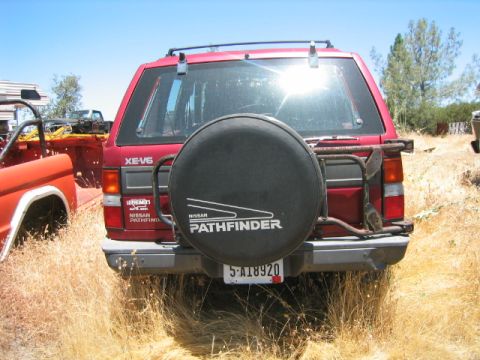 1973 Ford F-100 - A red Nissan Pathfinder is seen from the rear, parked in a dry, grassy area with another vehicle to its left.