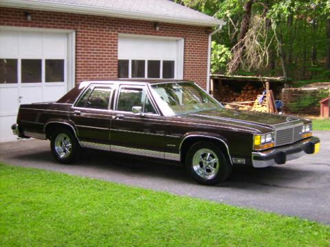 1982 Ford Crown Victoria LTD - A brown 1980s Ford LTD Crown Victoria is parked in front of a brick garage with a wooded area in the background.