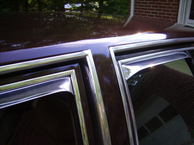 Dark car roof and chrome window trim with reflections of trees and brick house
