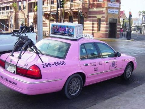2001 Ford Crown Victoria - A pink taxi cab with a bike rack is parked on a city street next to a building.