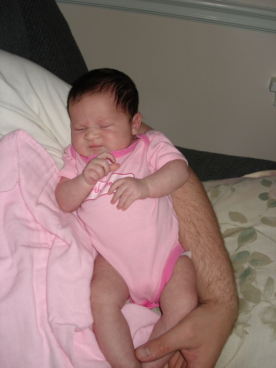 Newborn baby in pink onesie held by adult arms, with a pink blanket