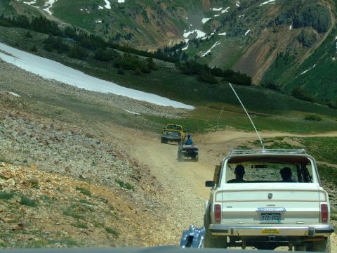 1987 Ford Bronco - Multiple off-road vehicles navigate a dusty mountain pass with snow patches visible on the slopes.