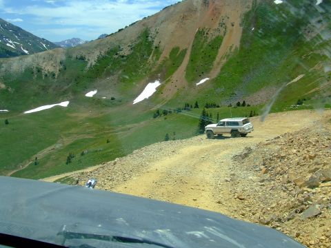 1987 Ford Bronco - A white SUV is parked on a dusty mountain road with green hills and snow patches in the background.