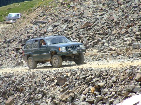 1987 Ford Bronco - A dark green Jeep Grand Cherokee navigates a challenging rocky terrain, with another vehicle following in the distance.