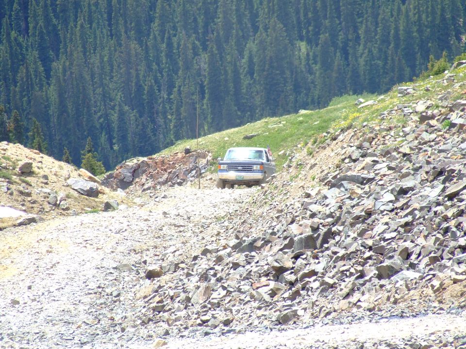 1987 Ford Bronco driving up a steep, rocky mountain road with pine trees in the background