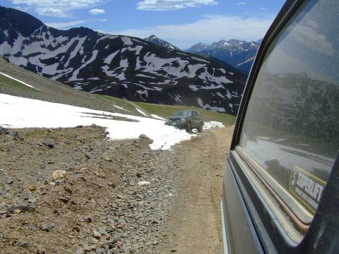 1987 Ford Bronco - A Jeep Cherokee navigates a snowy mountain pass, with another vehicle's perspective showing the terrain and distant peaks.