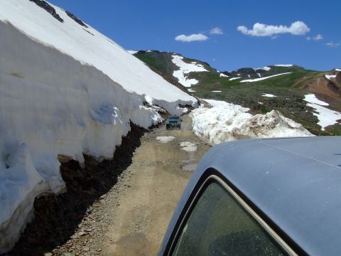 1987 Ford Bronco - Two SUVs navigate a narrow dirt road flanked by tall snowbanks under a bright blue sky.