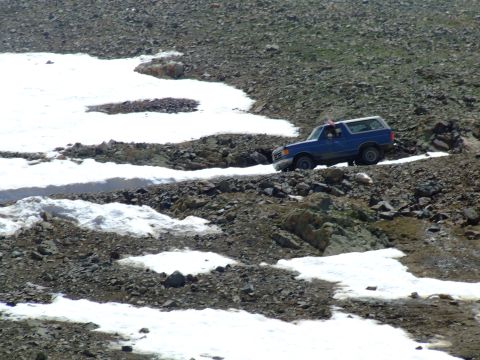 1987 Ford Bronco - A blue 1987 Ford Bronco navigates a challenging mountain trail with patches of snow and loose rocks.