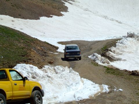 1987 Ford Bronco - Two vehicles navigate a snowy mountain road, with a yellow pickup truck in the foreground and a blue SUV ahead.