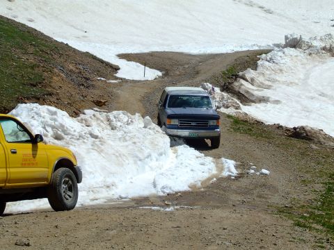 1987 Ford Bronco - A blue 1987 Ford Bronco drives through a snowy mountain pass, with a yellow truck visible in the foreground.
